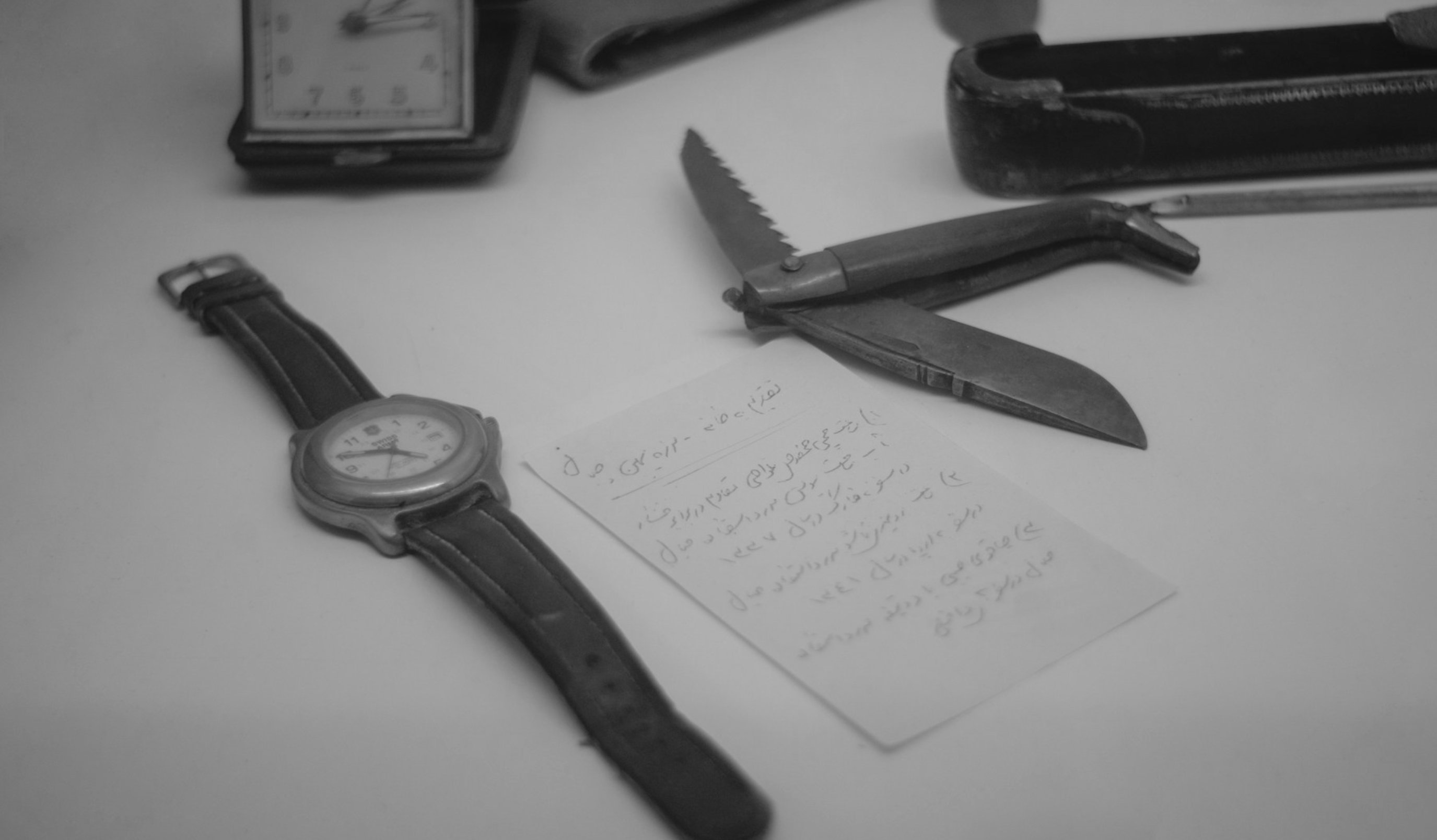 A wristwatch, a folding knife, a handwritten note and a travel clock on a table